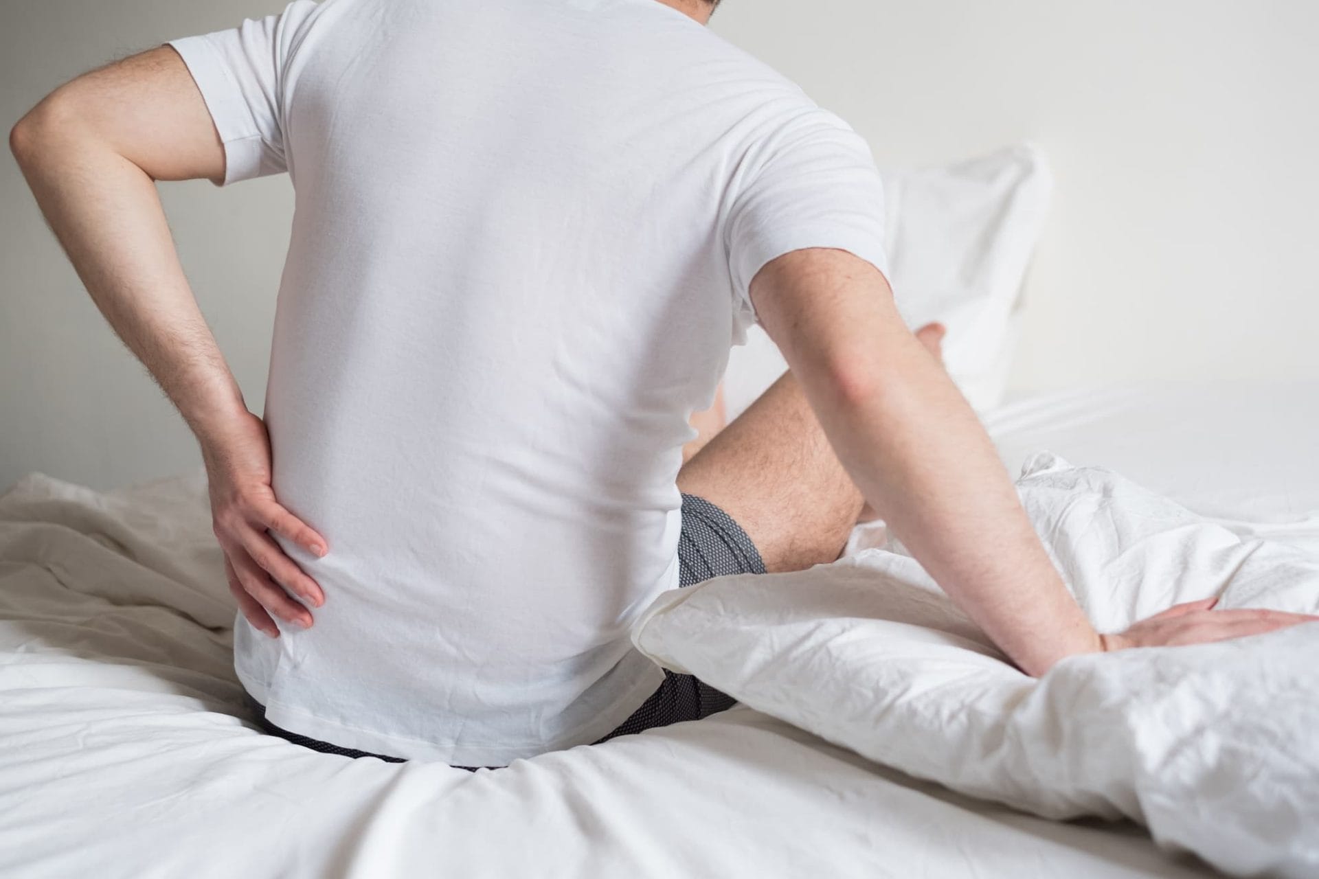 a man sat up right on the edge of his bed whilst holding his back in pain