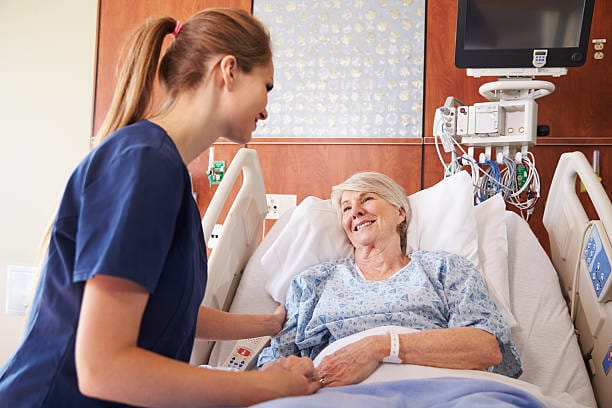 a patient sat up right in bed whilst smiling to a nurse who is checking on her